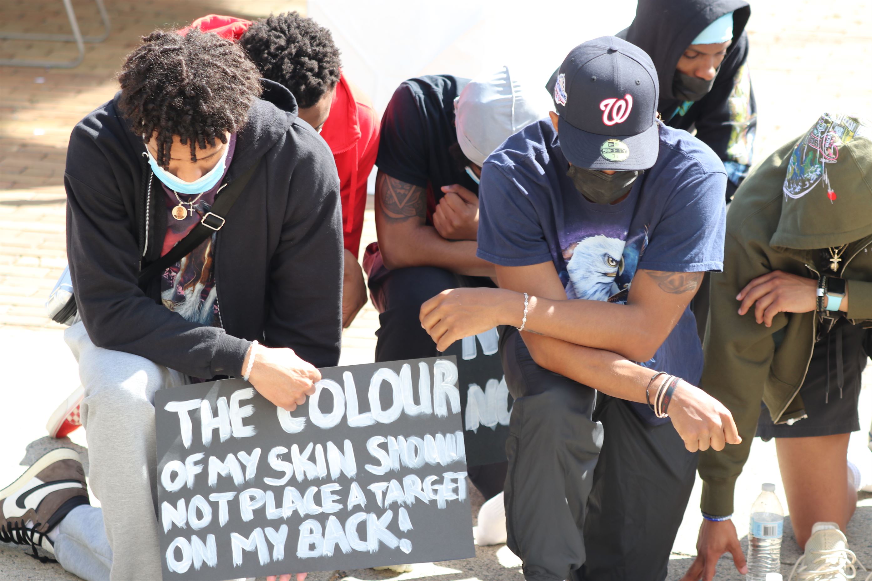 Black men take a knee for three minutes during a moment of silence, and one holds a poster that says “The colour of my skin should not place a target on my back!” Lynise Olivacce | The Montclarion