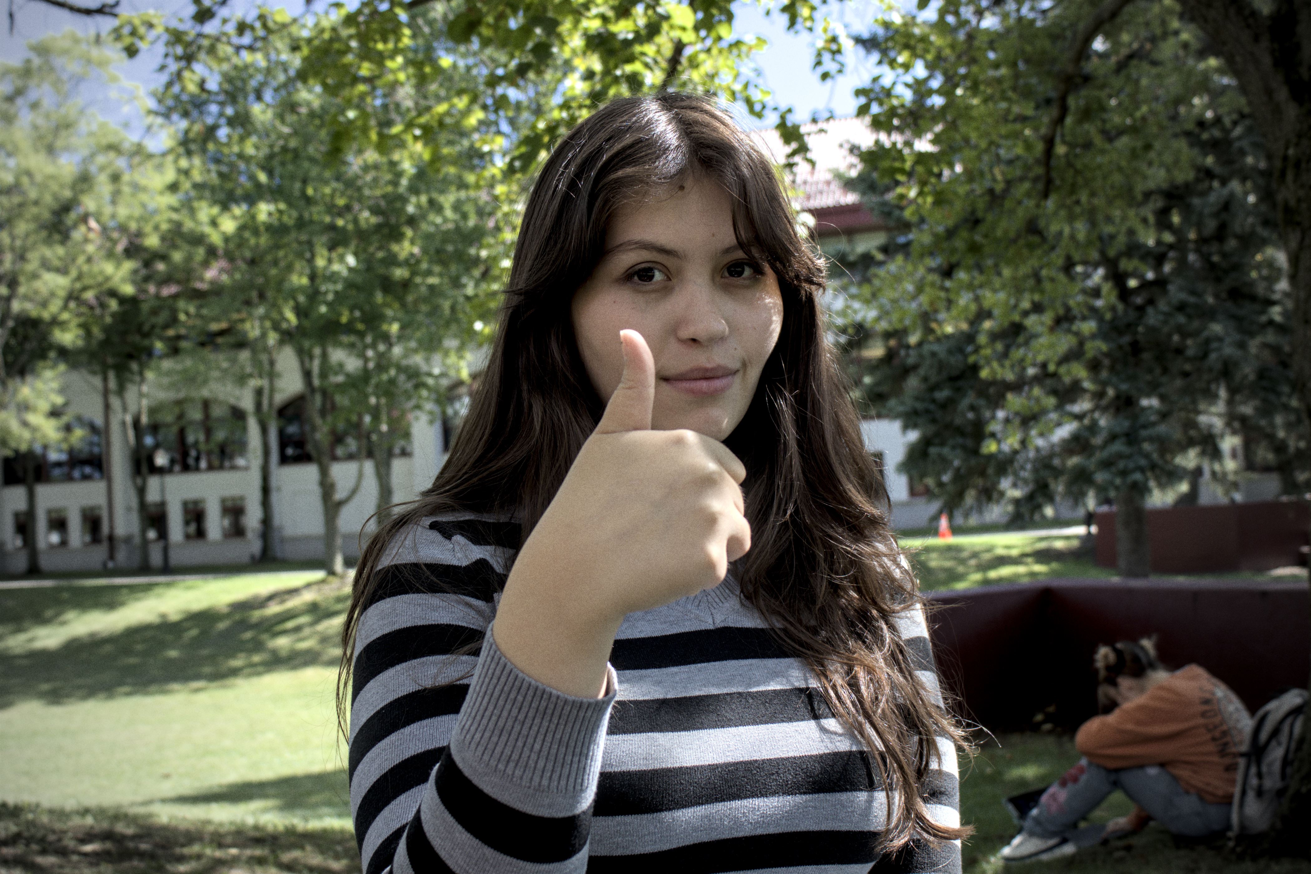Girl with brown hair down past her shoulders poses with a thumbs up. She is wearing a gray and black striped shirt.