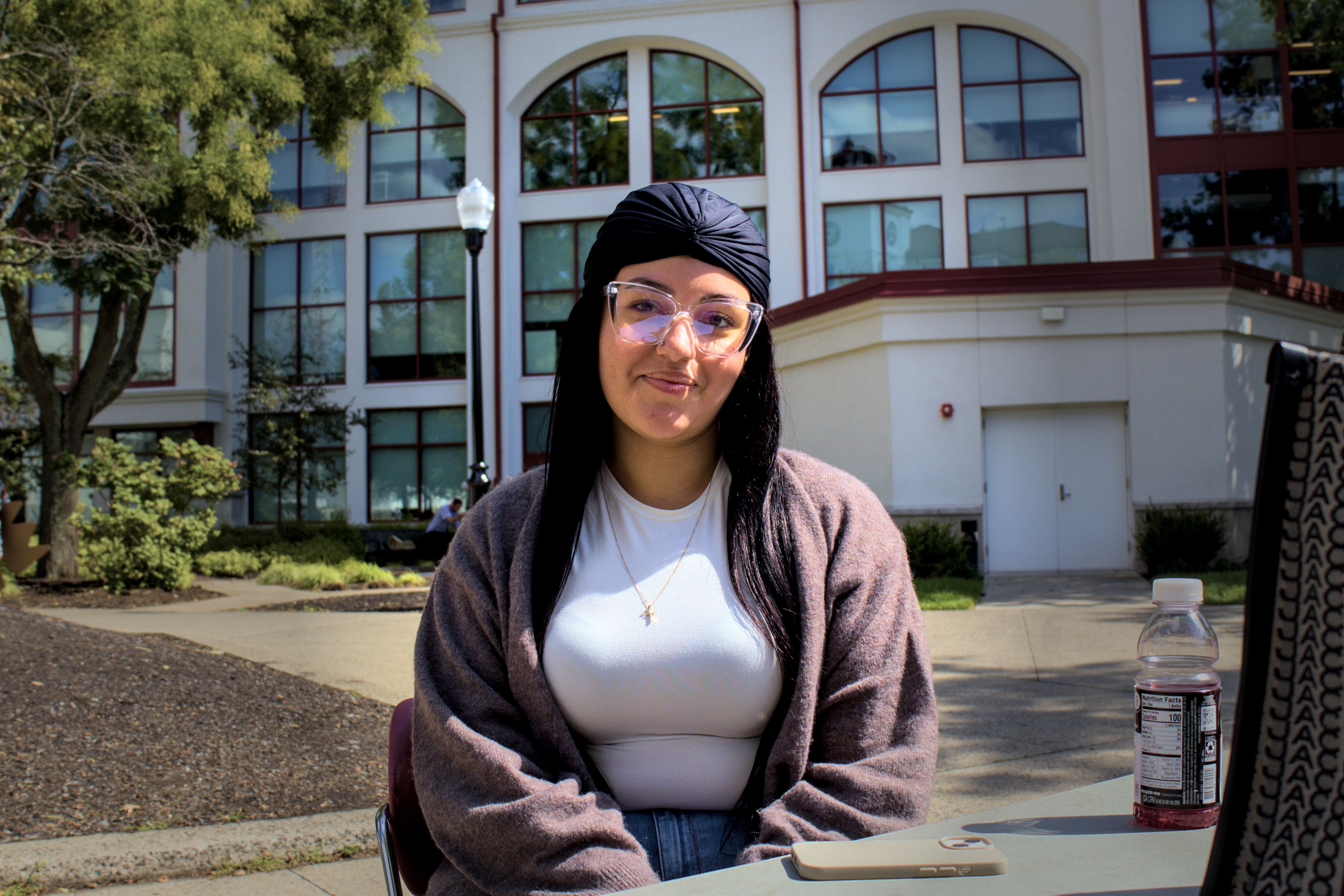 Girl with a blue headwrap and clear glasses sits in front of a building. Her black hair goes down to her chest. She is wearing a white shirt with blue jeans, covered by a gray sweater.