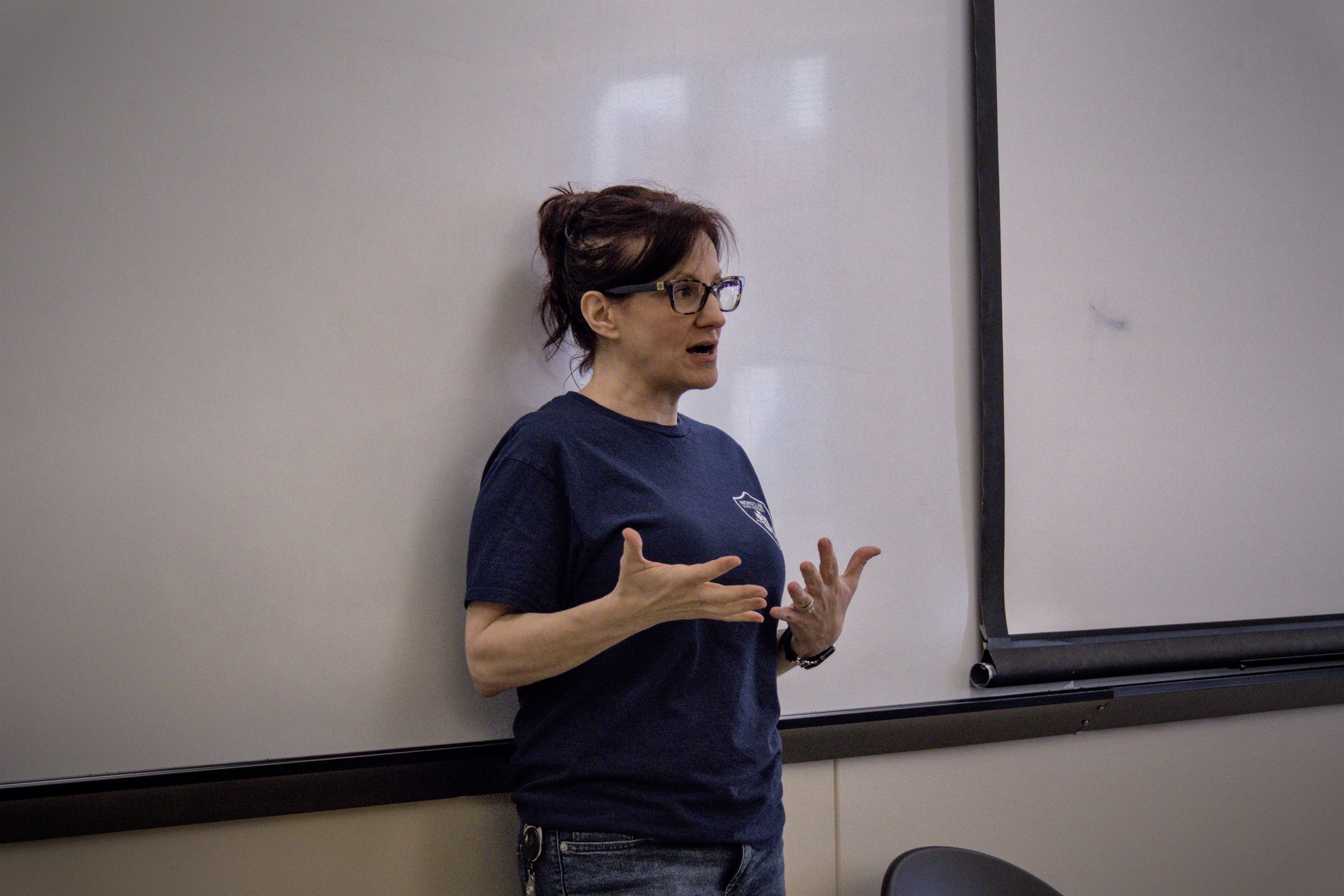 Red-headed woman with brown glasses explains concept to students. She is wearing a blue EMS shirt and blue jeans. Her hands are open to demonstrate while she is explaining.
