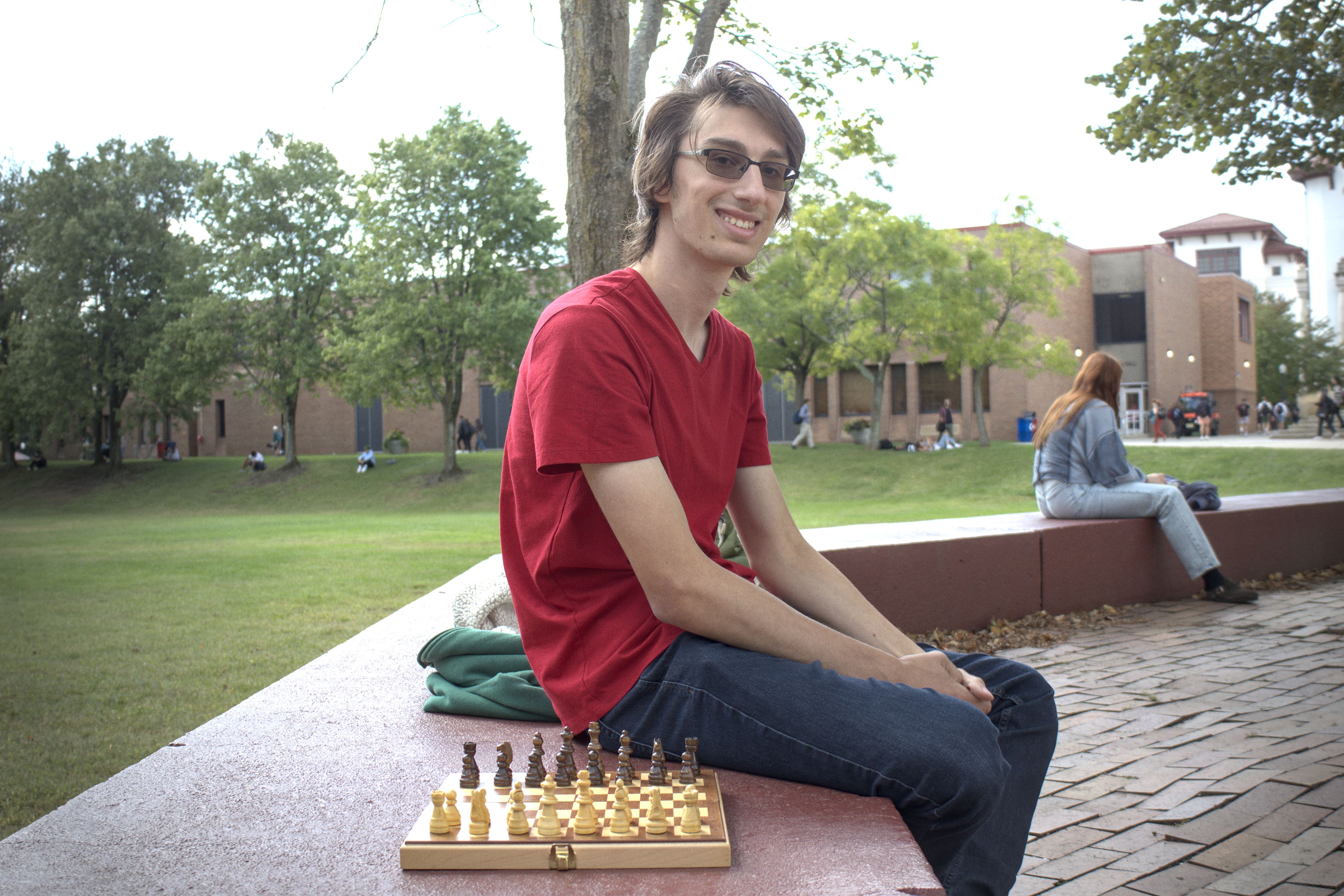 Tall student with long brown hair sits on maroon wall. His glasses are tinted, but do not obscure his eyes. Student is wearing a red t-shirt with blue jeans and is behind a portable chess set.