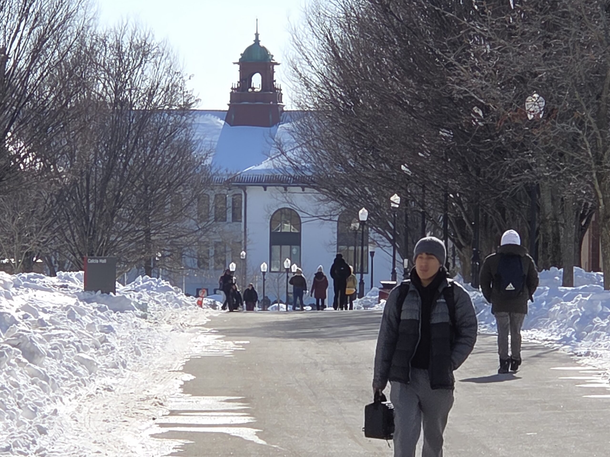 Students walk along a snowbank flanked  University Promenade with Cole Hall in the background.